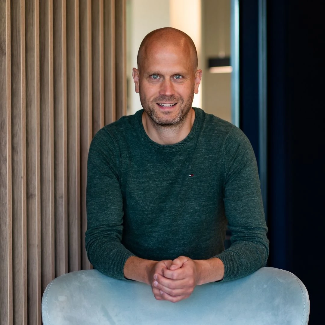 Tom van Stokkem heeft een donker groene trui aan en leunt op een witte stoel die voor een houten muur staat terwijl hij denkt aan de contramal van EPS die hij moet tekenen.