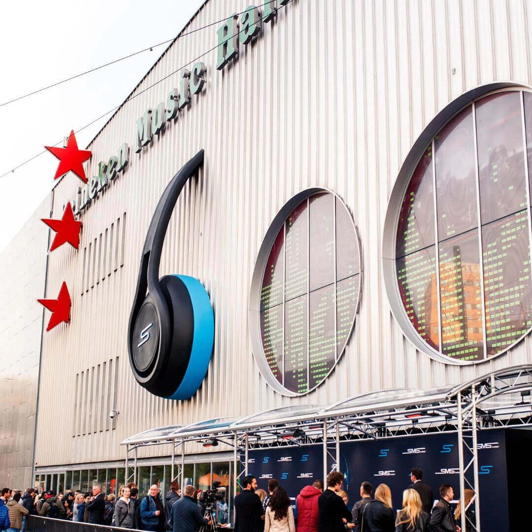De buitenkant van Heineken music hall met daarop een grote koptelefoon met zwarten en blauwe aspecten en de drie roden sterren van Heineken.