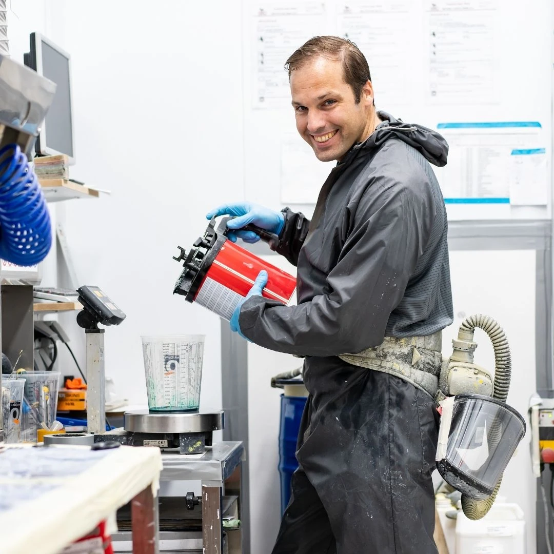 Twan van de Meulengraaf kijkt lachend in de camera terwijl hij, gehuld in beschermende kleding, het spuitreservoir vult met polyureacoating waarmee hij een piepschuim kunstwerk gaat afwerken..